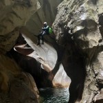 Eine Person, die in voller Canyoning-Ausrüstung an einer felsigen Wand sitzt, während Wasser unter ihr fließt.
